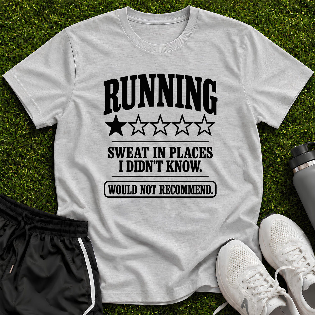 A light gray graphic t-shirt laid flat on green grass, featuring the text 'RUNNING SWEAT IN PLACES I DIDN'T KNOW. WOULD NOT RECOMMEND.
