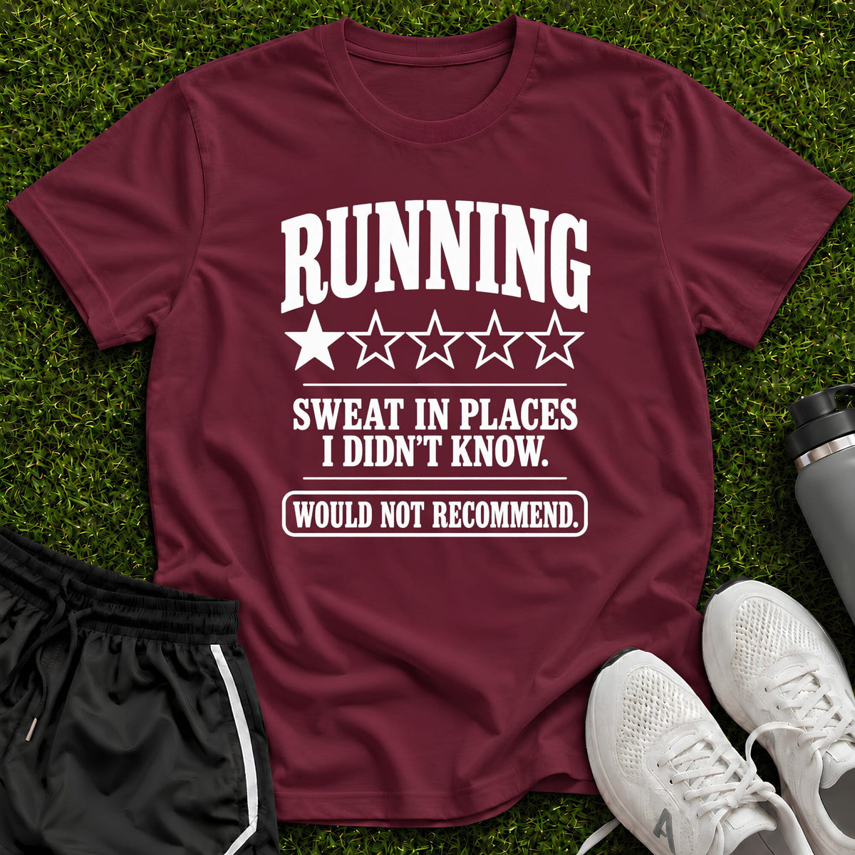 A maroon short-sleeve graphic T-shirt laid flat on green grass, with white text reading “RUNNING SWEAT IN PLACES I DIDN'T KNOW. WOULD NOT RECOMMEND.