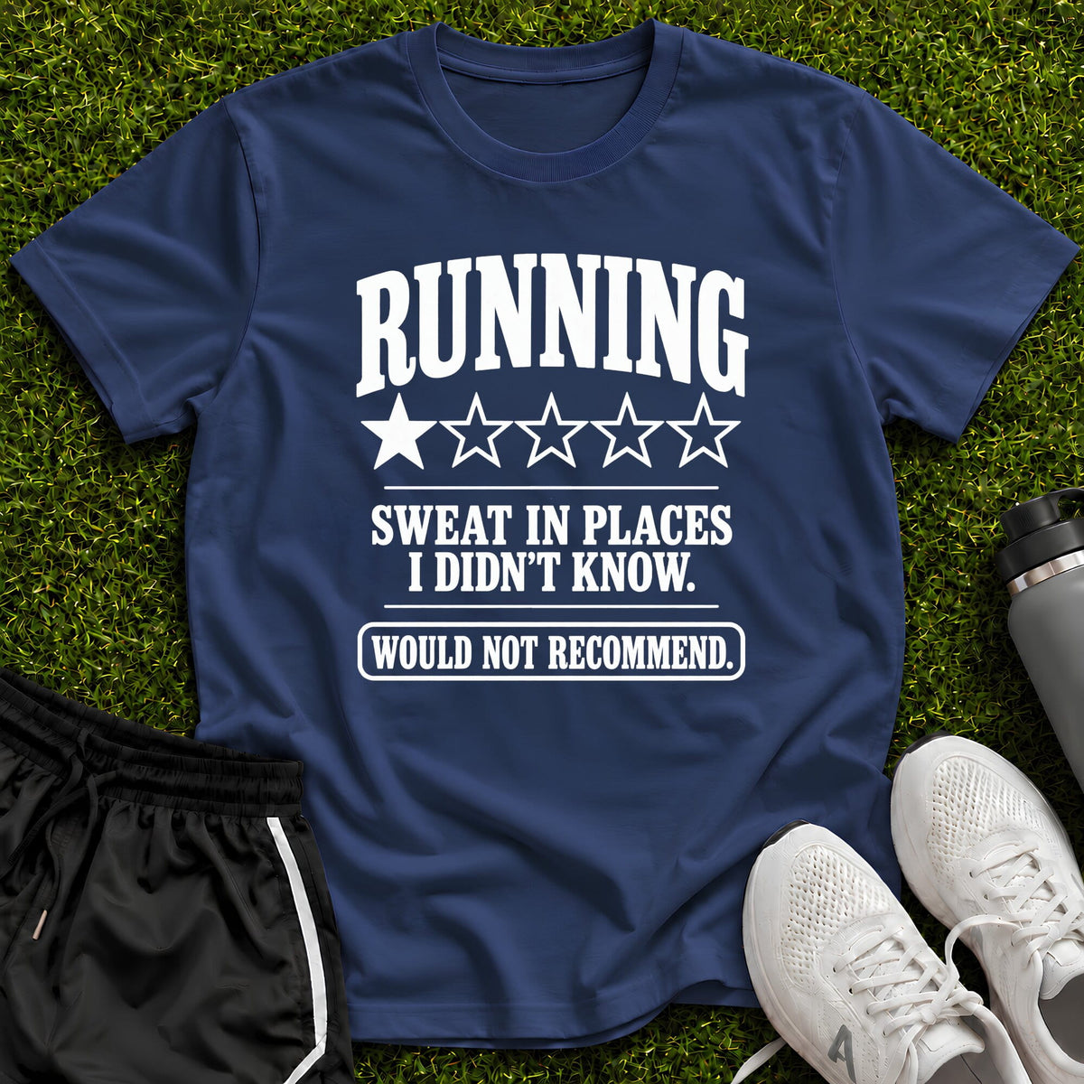 A navy blue graphic t-shirt laid flat on green grass, styled with black running shorts, white sneakers, and a water bottle. The t-shirt features a white graphic that reads 'RUNNING SWEAT IN PLACES I DIDN'T KNOW.